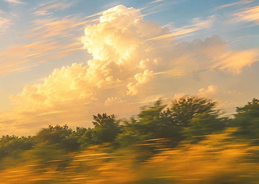 Towering golden cumulus clouds at sunset above a treeline and a field of grass blurred by motion.